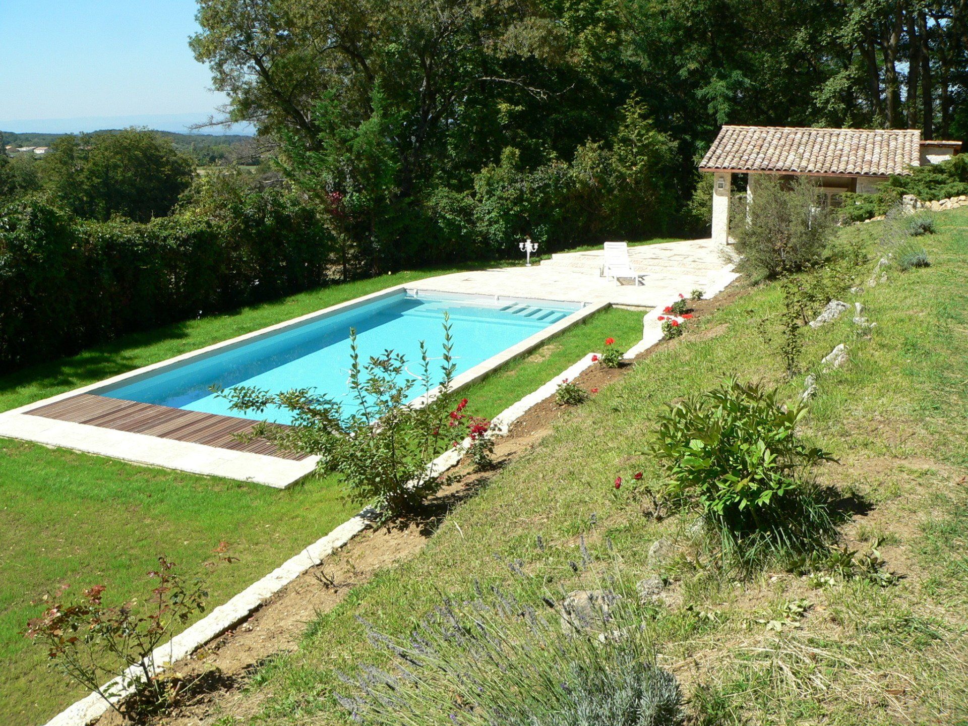 Piscine rectangulaire dans un jardin gazonné, avec un petit bâtiment et des arbres environnants.