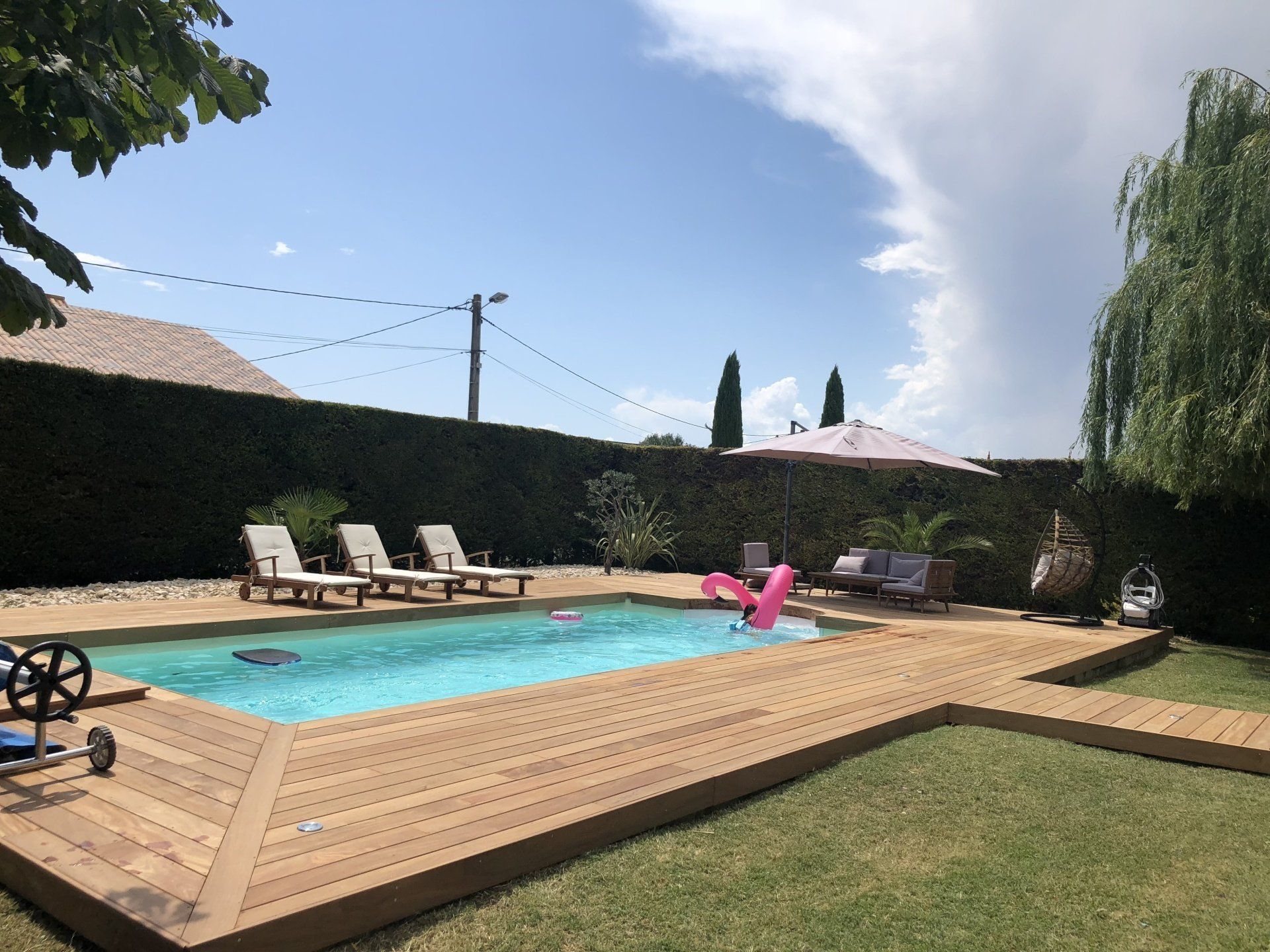 Piscine aux formes arrondies, entourée d'une zone pavée, de pelouse et d'arbres sous un ciel ensoleillé.