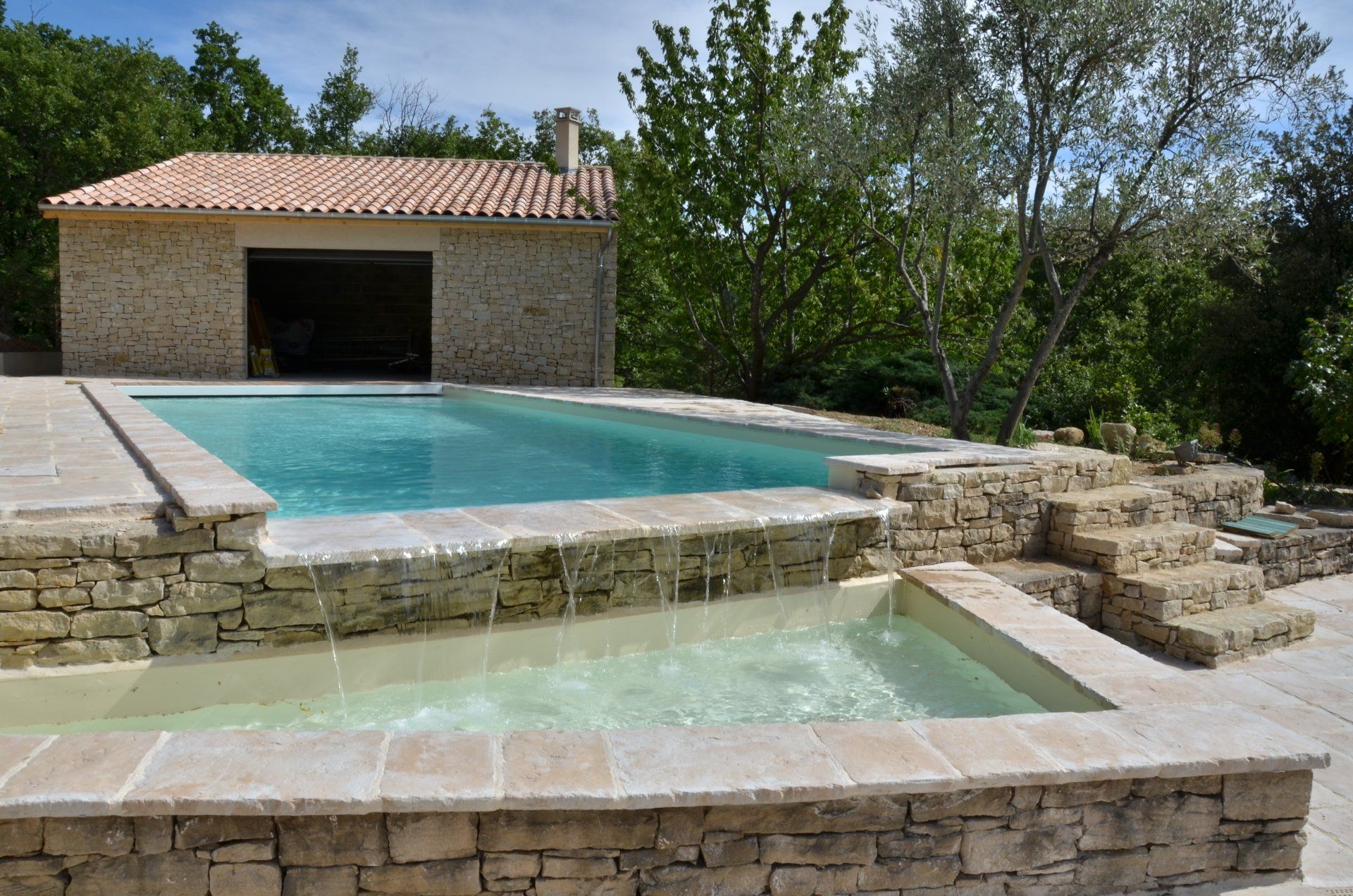 Une piscine rectangulaire avec un niveau inférieur, de l'eau courante, des murs en pierre et un petit bâtiment dans un cadre extérieur ensoleillé.