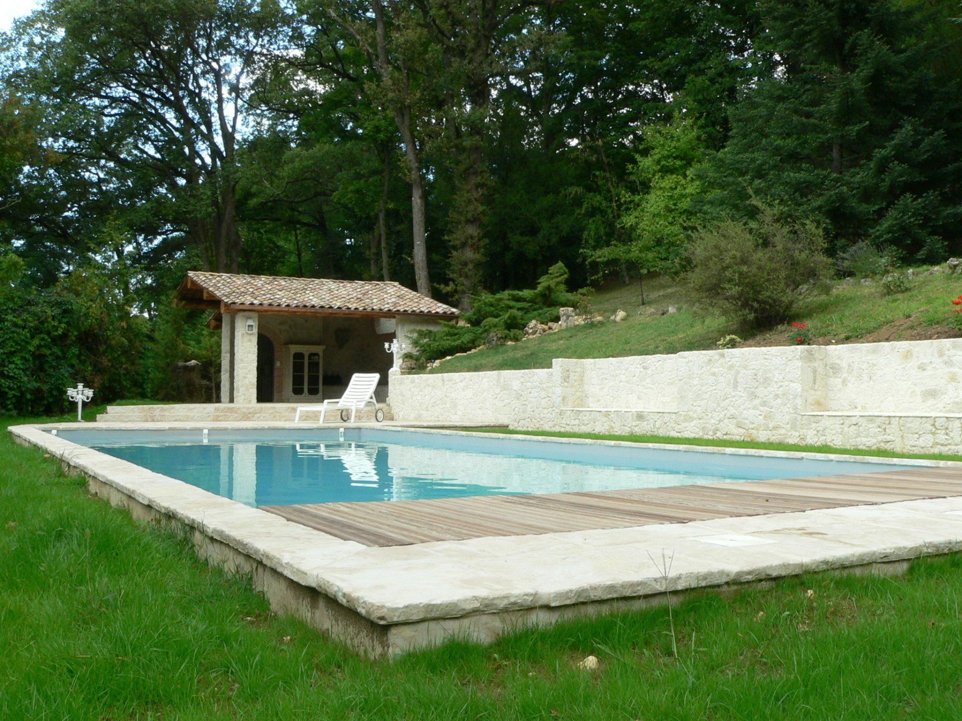 Piscine entourée de pierres, jardin gazonné et petit bâtiment en pierre dans un cadre boisé.