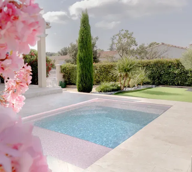 Une piscine extérieure rectangulaire avec des marches, entourée de pierres claires, de pelouse verte et offrant une vue sur une colline au loin.