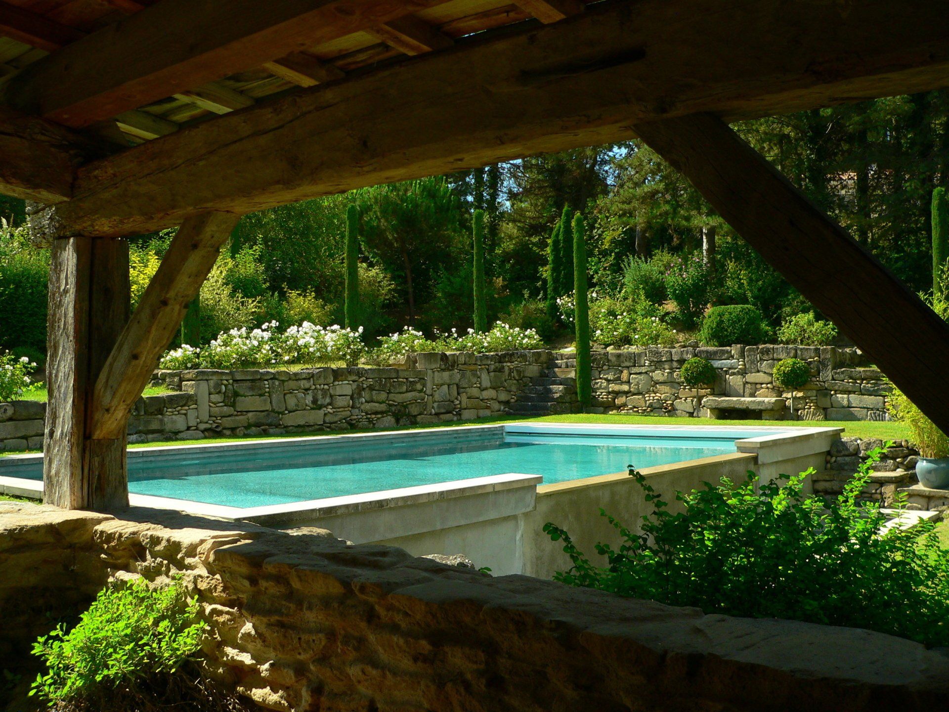 Piscine vue depuis une structure en bois ombragée, entourée de murs en pierre et d'une végétation luxuriante.