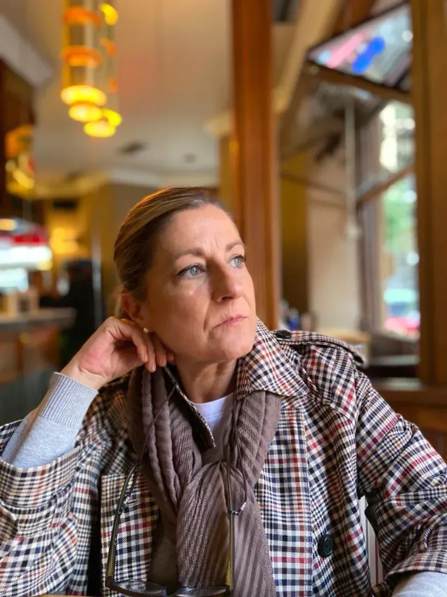 Mujer con abrigo a cuadros mirando por la ventana de un café, apoyando la cabeza en su mano.