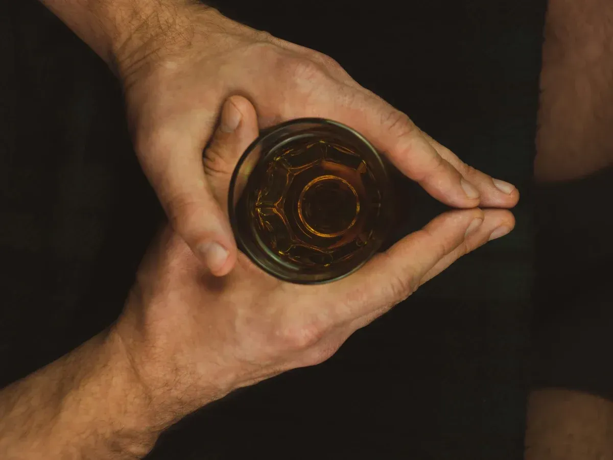 Manos sosteniendo un vaso de líquido ámbar, vista de arriba hacia abajo, sobre un fondo negro.