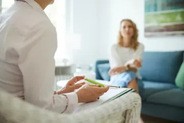 Terapeuta con portapapeles y bolígrafo, hablando con una mujer en un sofá azul en una habitación muy iluminada.