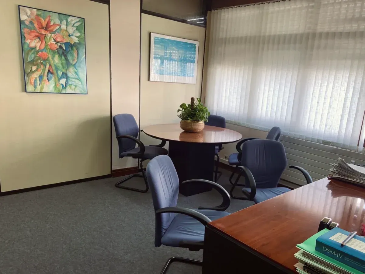 Área de reuniones de oficina: mesa redonda, sillas azules, obras de arte, planta en maceta y ventana con persianas.