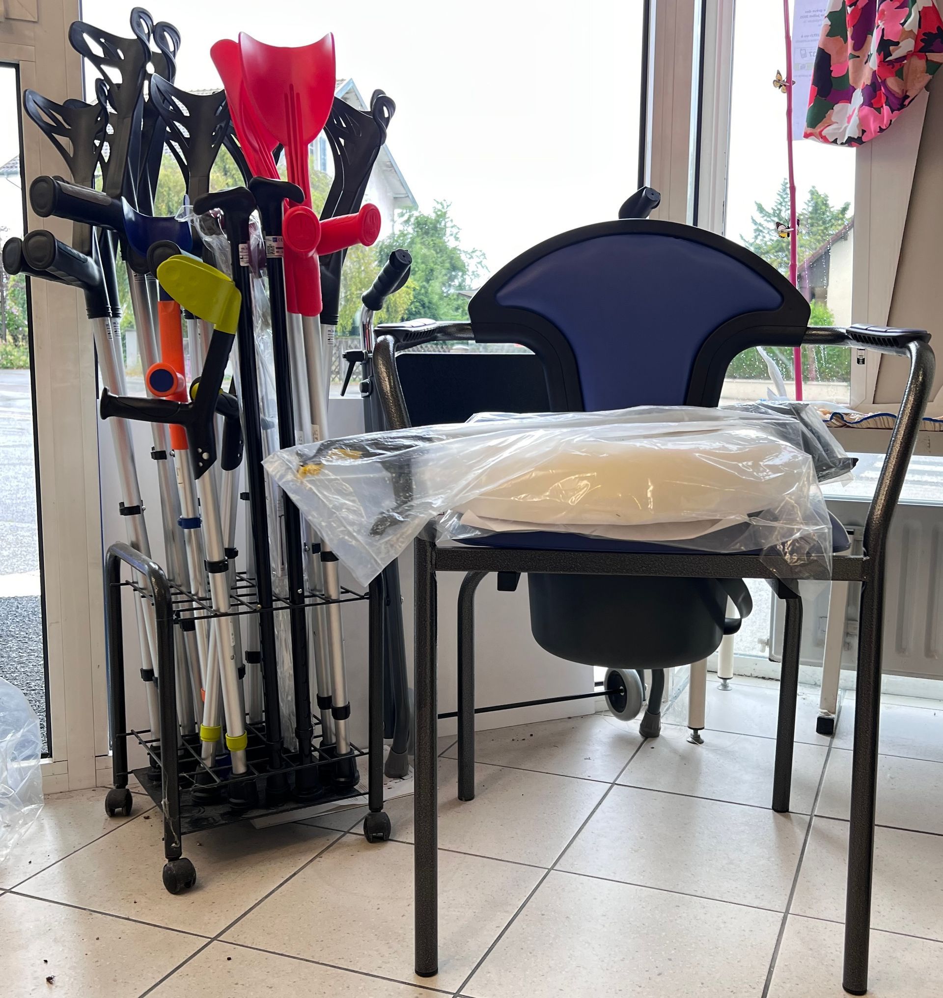 Des aides à la mobilité pliées sont placées à côté d'une chaise dans une pièce carrelée et lumineuse, avec une table recouverte d'un drap au premier plan.