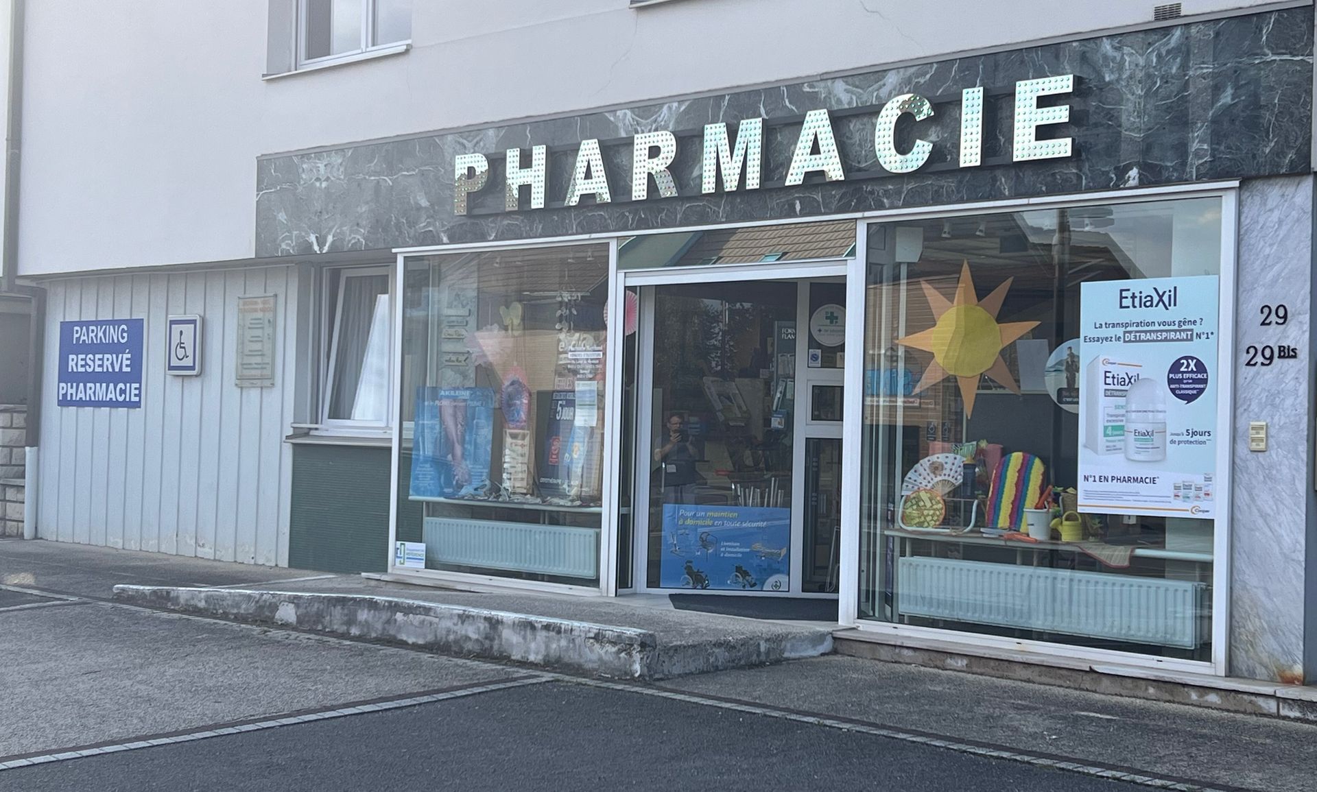 Façade de pharmacie avec entrée vitrée, grande enseigne PHARMACIE et affiches sur les vitrines.
