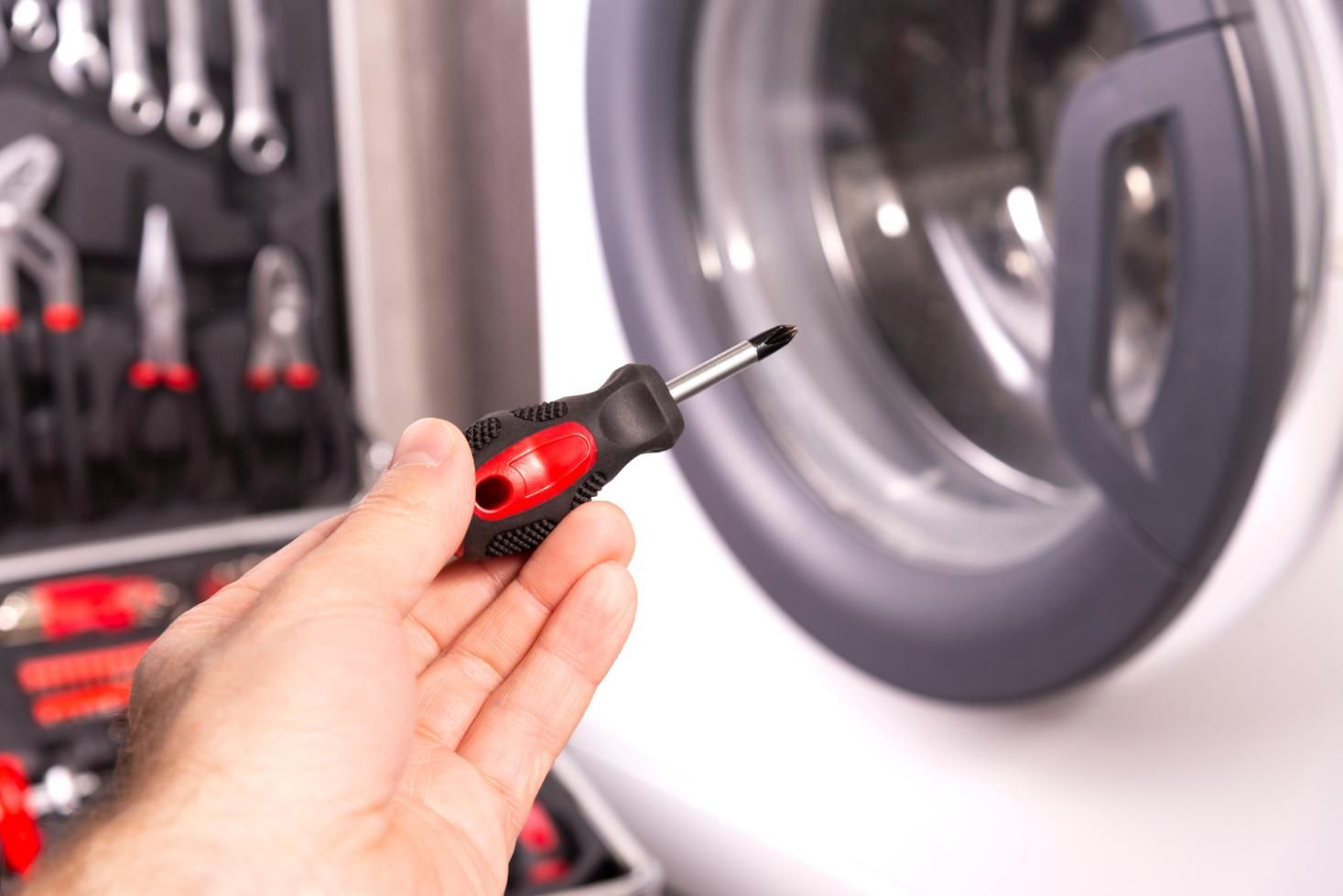 Mano sosteniendo un destornillador cerca de una lavadora; herramientas de reparación en el fondo.