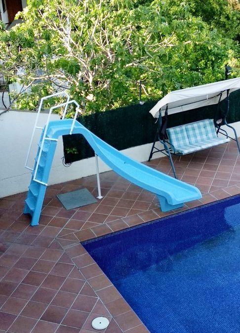 Tobogán de piscina azul, columpio y piscina en un patio de baldosas.