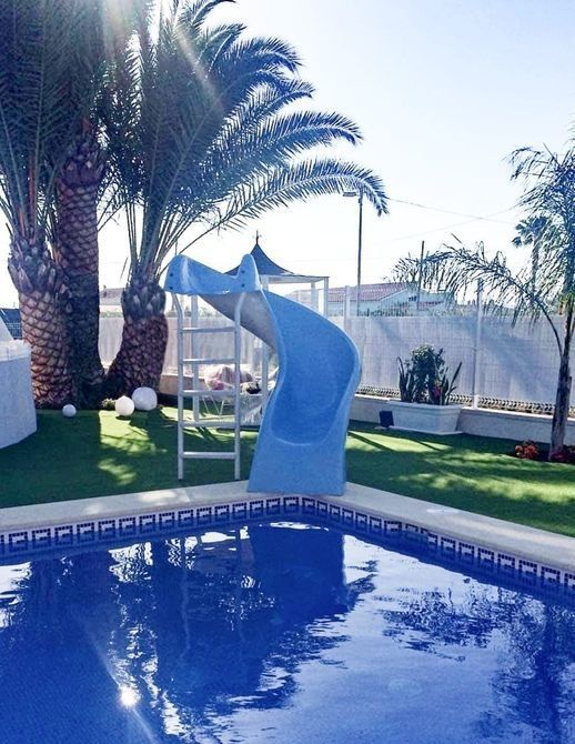 Un tobogán de agua azul desciende hacia una piscina de azulejos azules rodeada de césped verde y palmeras en un día soleado.