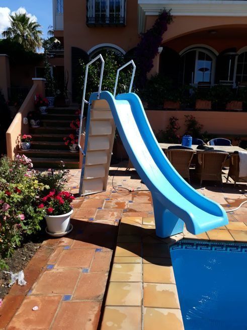 Tobogán de piscina azul junto a una piscina con escaleras y una casa al fondo.