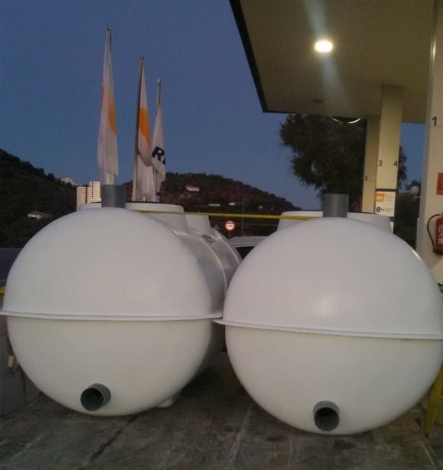 Tanques esféricos blancos en una gasolinera, con tuberías y banderas en el fondo.