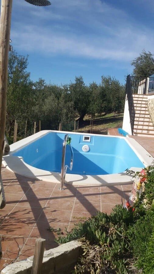 Piscina azul vacía con ducha y patio de ladrillo, rodeada de árboles bajo un cielo azul.
