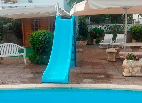 Tobogán azul junto a una piscina. Patio con bancos, mesa y una pequeña estructura al fondo.