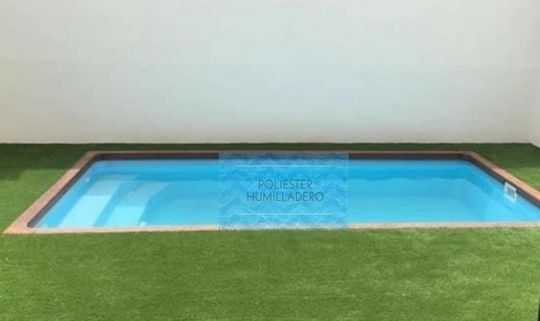 Piscina rectangular de color azul, rodeada de césped artificial verde y borde de madera, frente a una pared blanca.