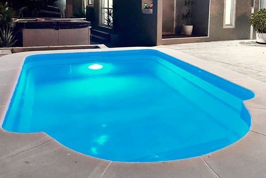 Piscina con iluminación azul y esquinas redondeadas. Patio de hormigón alrededor.