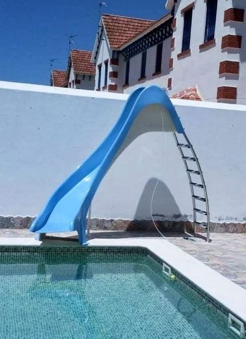 Tobogán de agua azul sobre una piscina, pared blanca, edificios al fondo.