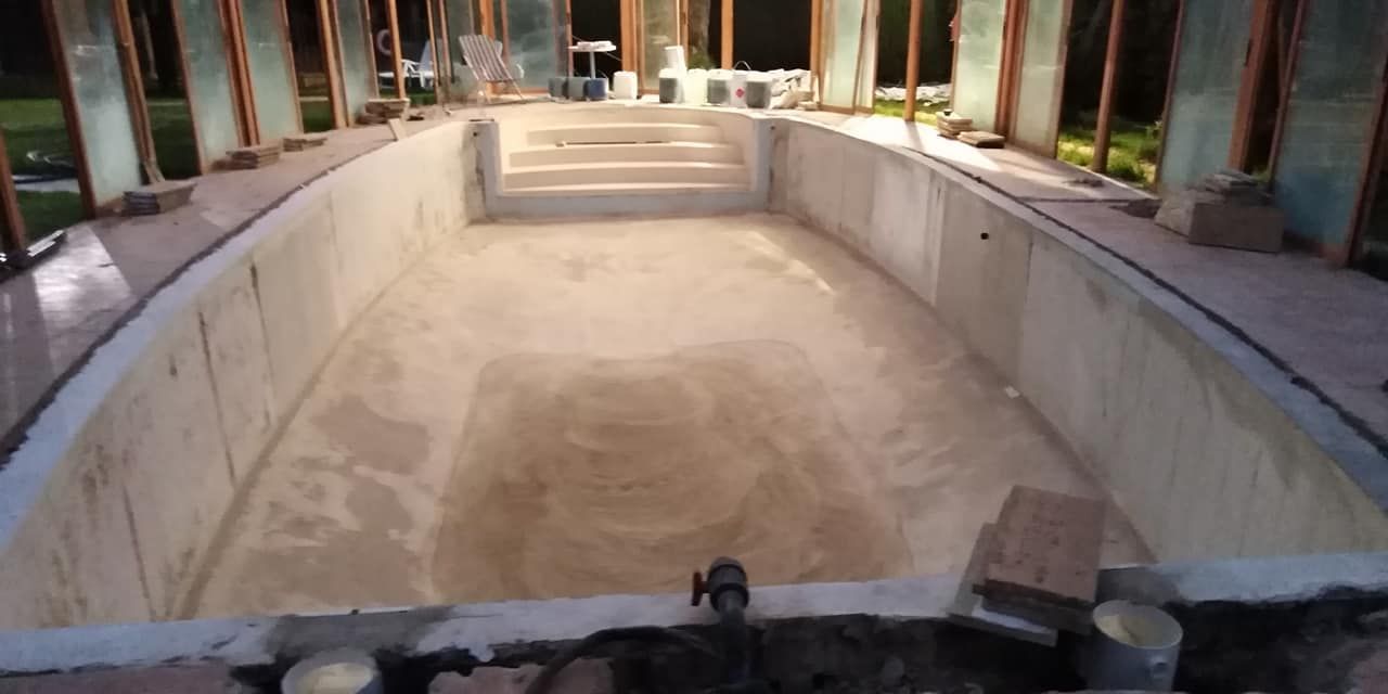 Piscina de hormigón vacía en construcción, con escalones en la parte menos profunda, dentro de una estructura con paredes de vidrio.