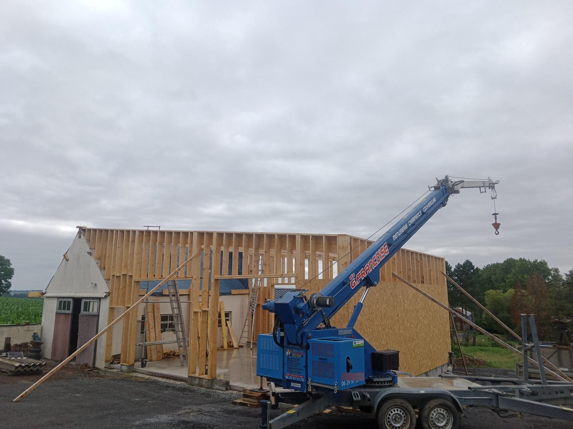 Une petite grue bleue soulève des poutres en bois pour un bâtiment en construction par temps nuageux.