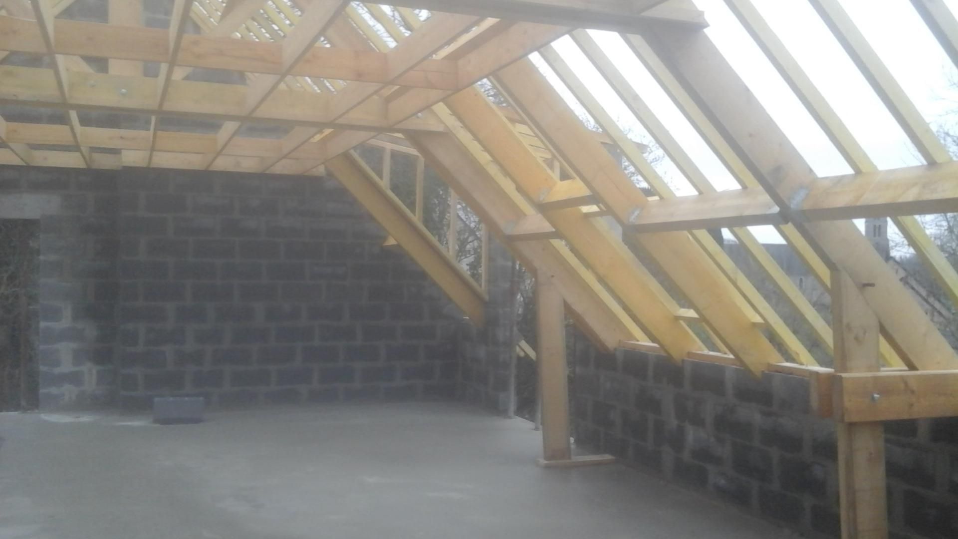Intérieur d'un bâtiment en construction, montrant une charpente de toit en bois et des murs en parpaings.