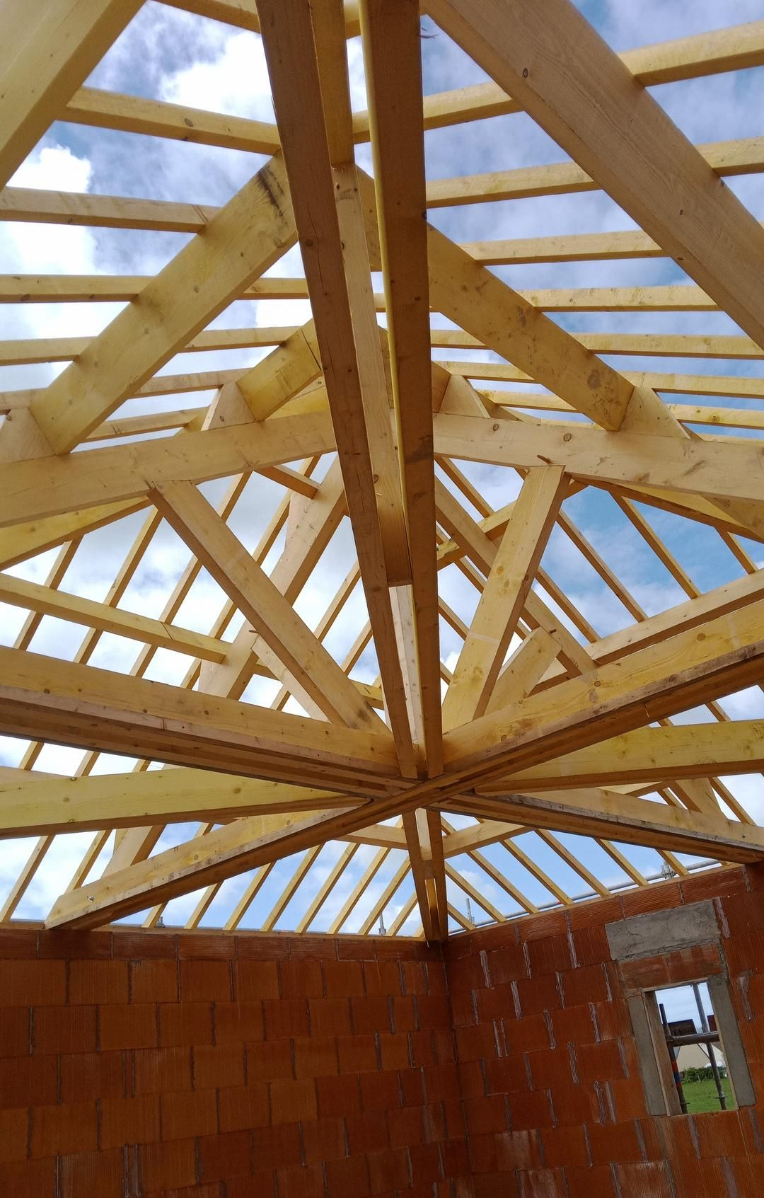 Charpente de toit en bois en construction sous un ciel partiellement nuageux. Murs en briques rouges visibles.