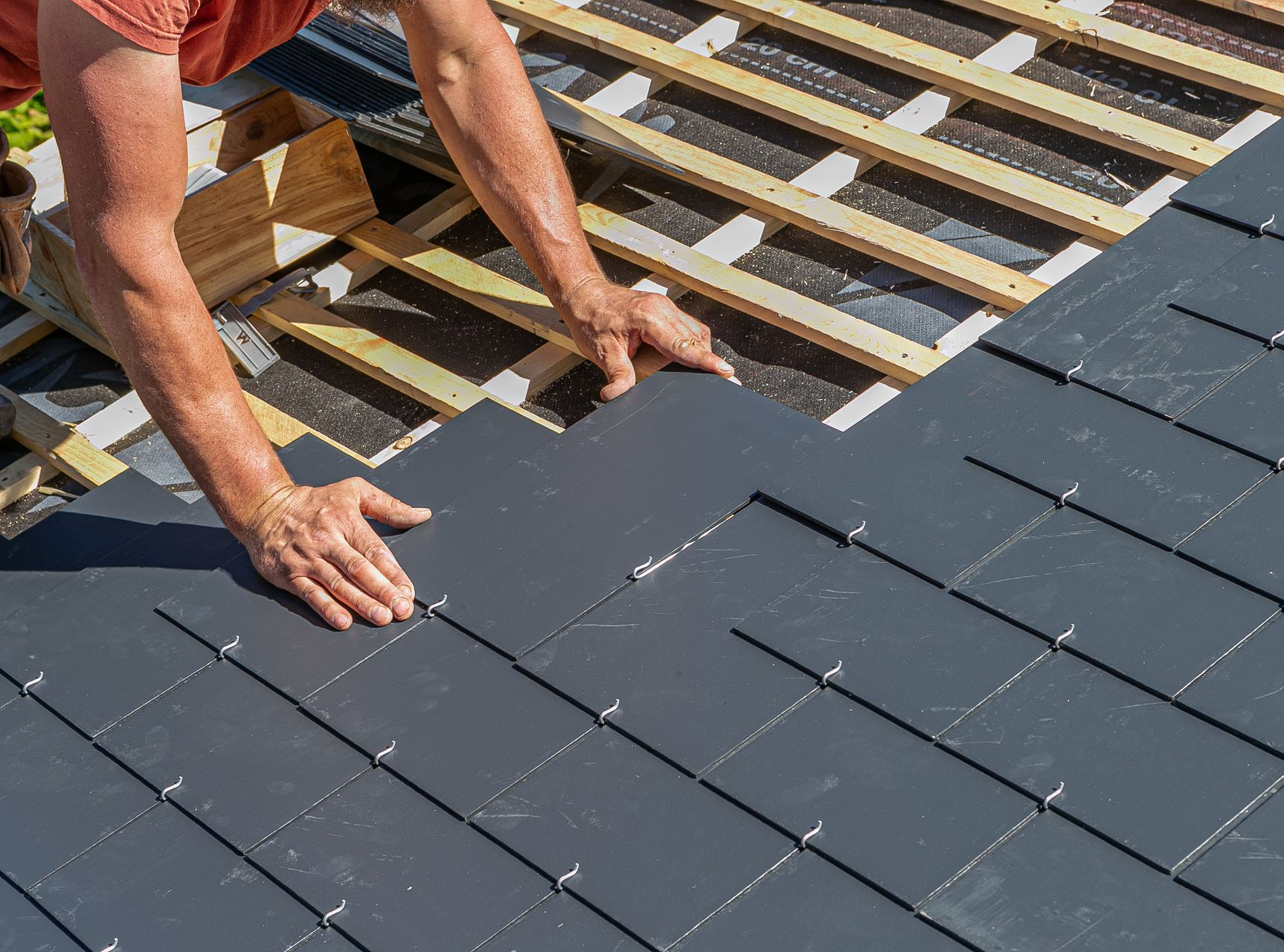 Une personne installe des tuiles de toit gris foncé sur une charpente en bois.