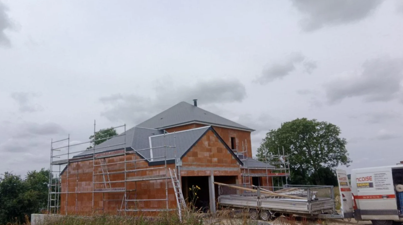 Maison en construction, façade en briques rouges, échafaudages, toit gris, ciel couvert.