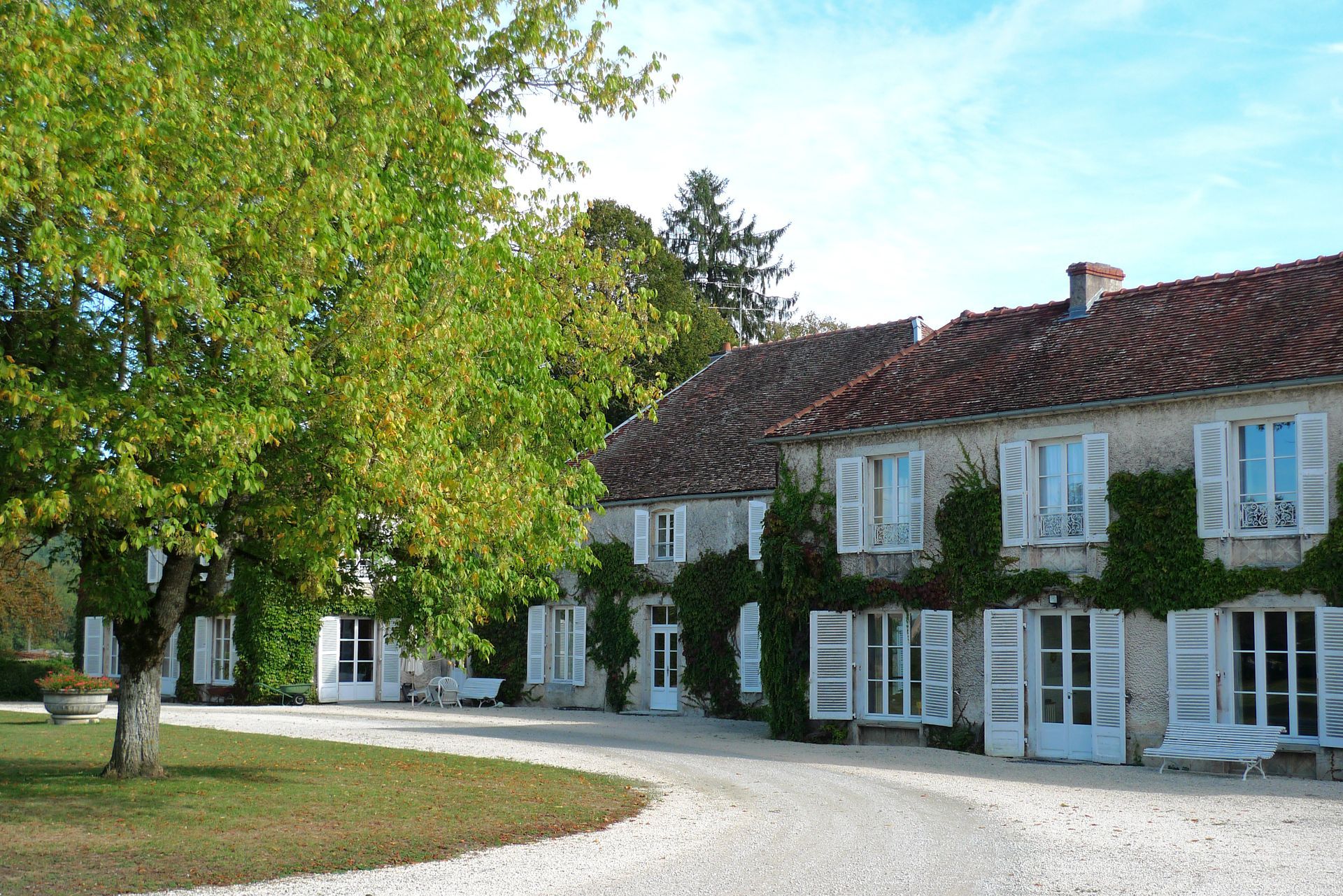 Maison de campagne aux volets blancs avec du lierre sur la façade, située à côté d'une allée de gravier.
