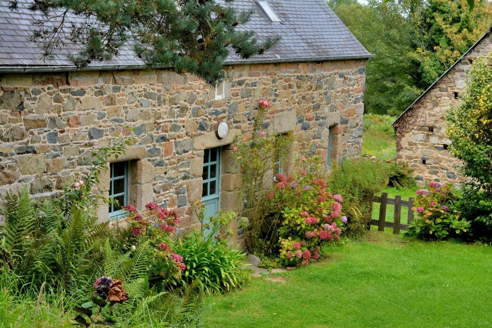 Maisonnette en pierre ornée de lierre et de fleurs roses, bordée d'une pelouse verdoyante.