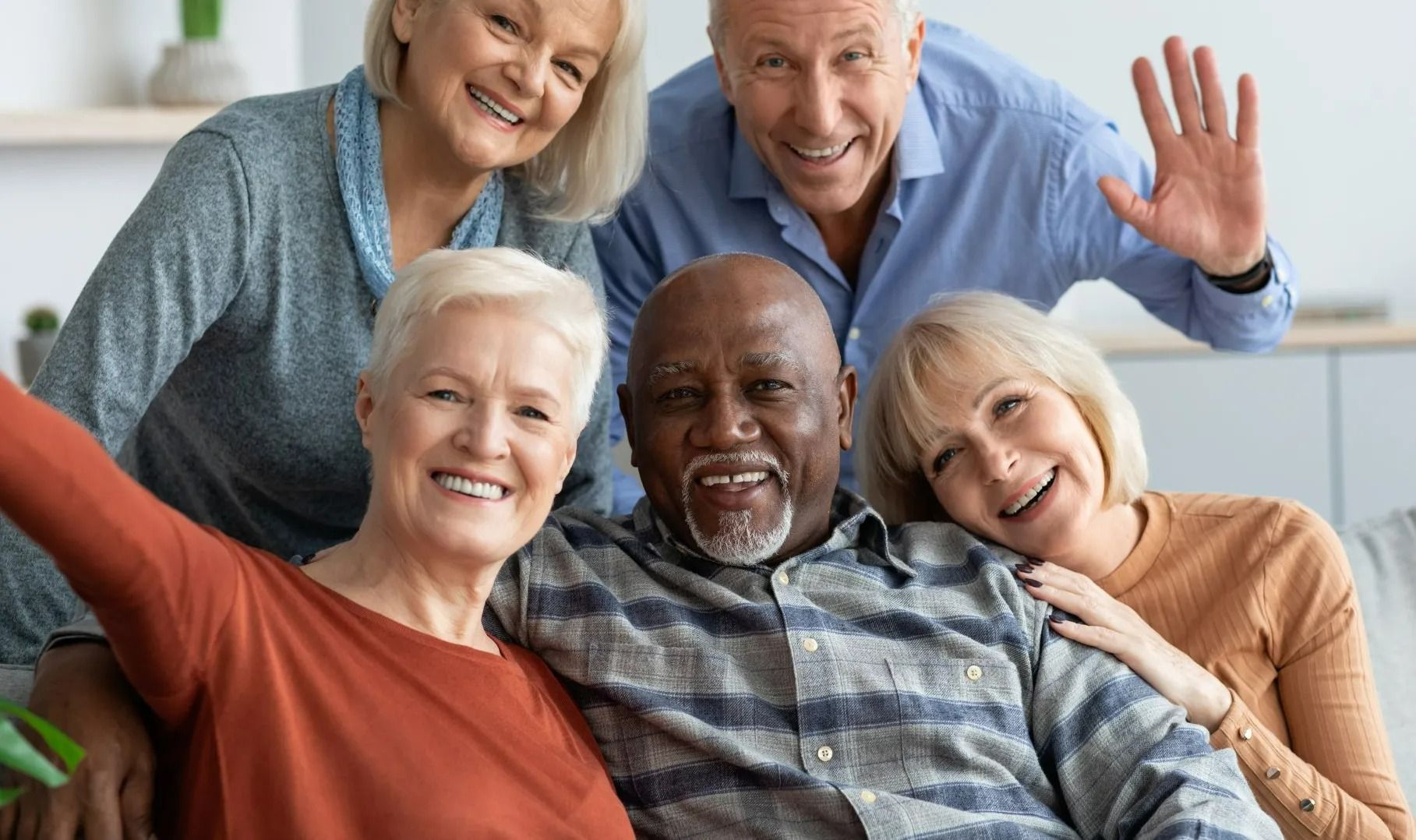 Cinq adultes souriants, assis ensemble sur un canapé, font signe à la caméra.