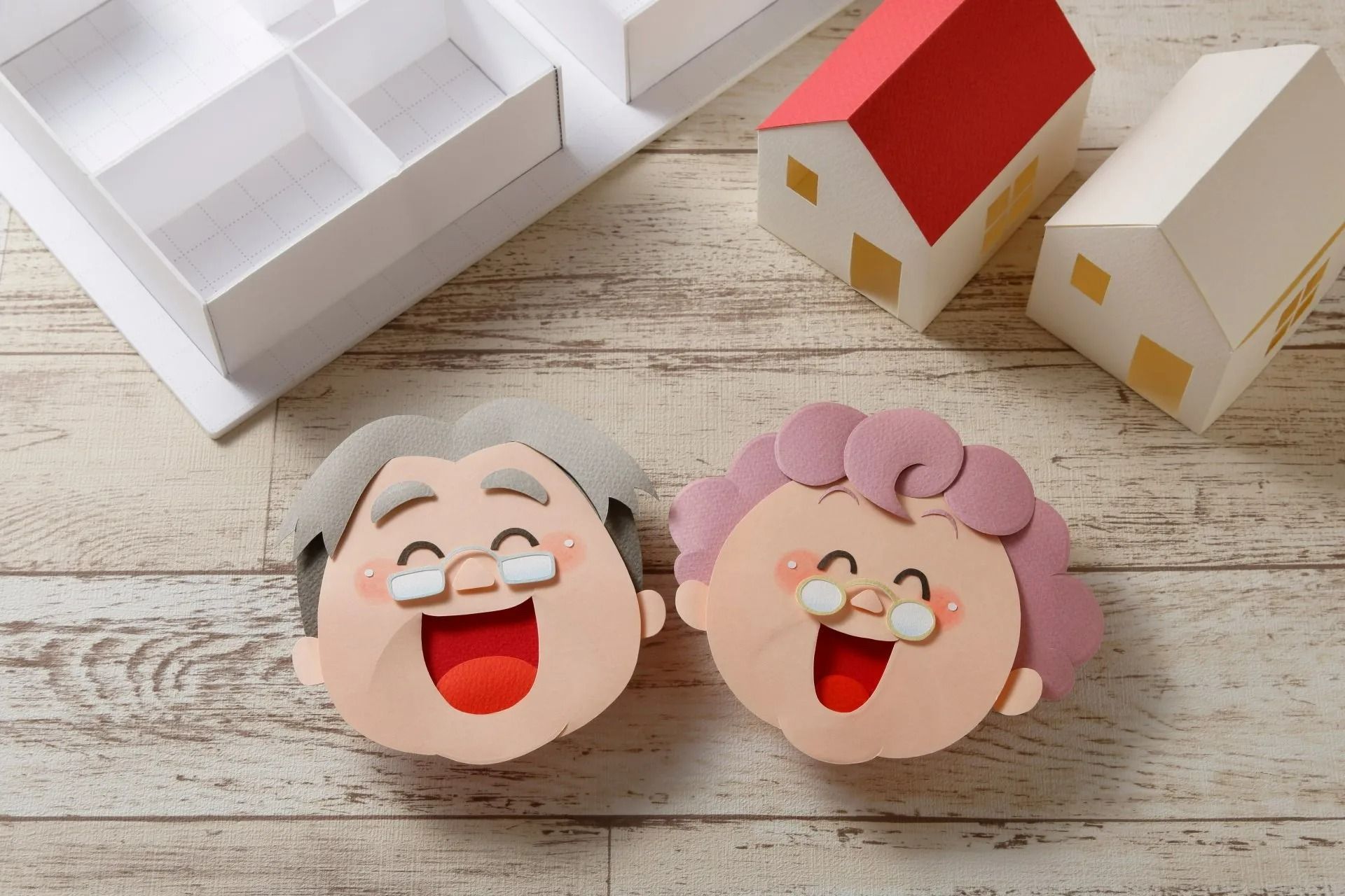 Deux visages souriants en papier sur une table, à proximité de petites maquettes de maisons.