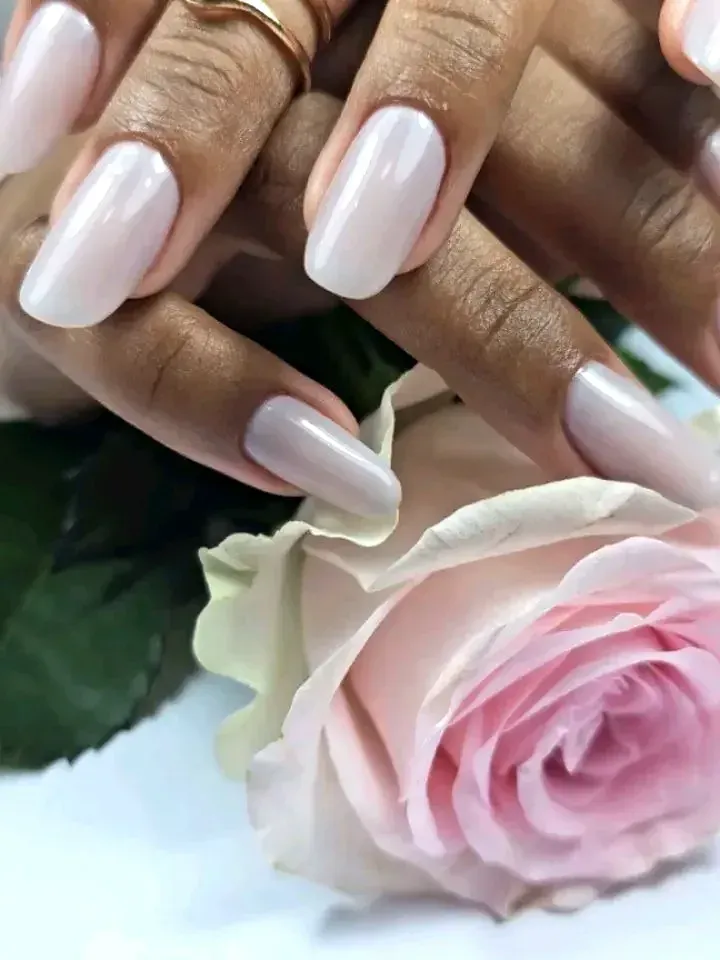 Manos con uñas cuidadas de color rosa pálido y acabado brillante junto a una rosa rosa