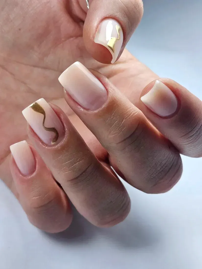 Mano con uñas largas color nude y detalles dorados en espiral sobre un fondo azul pálido