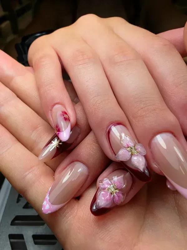 Manos con uñas acrílicas largas en tonos nude y rosa con motivos florales; un dedo lleva un charm plateado.