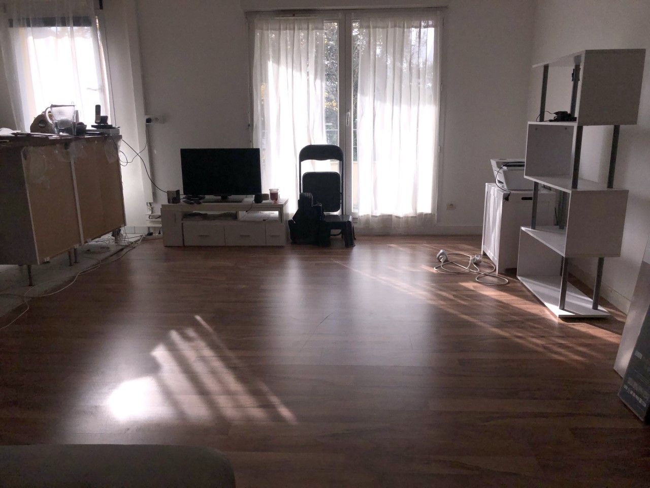 Un salon lumineux avec un parquet foncé, une télévision sur un meuble, un fauteuil noir et une étagère blanche ouverte.