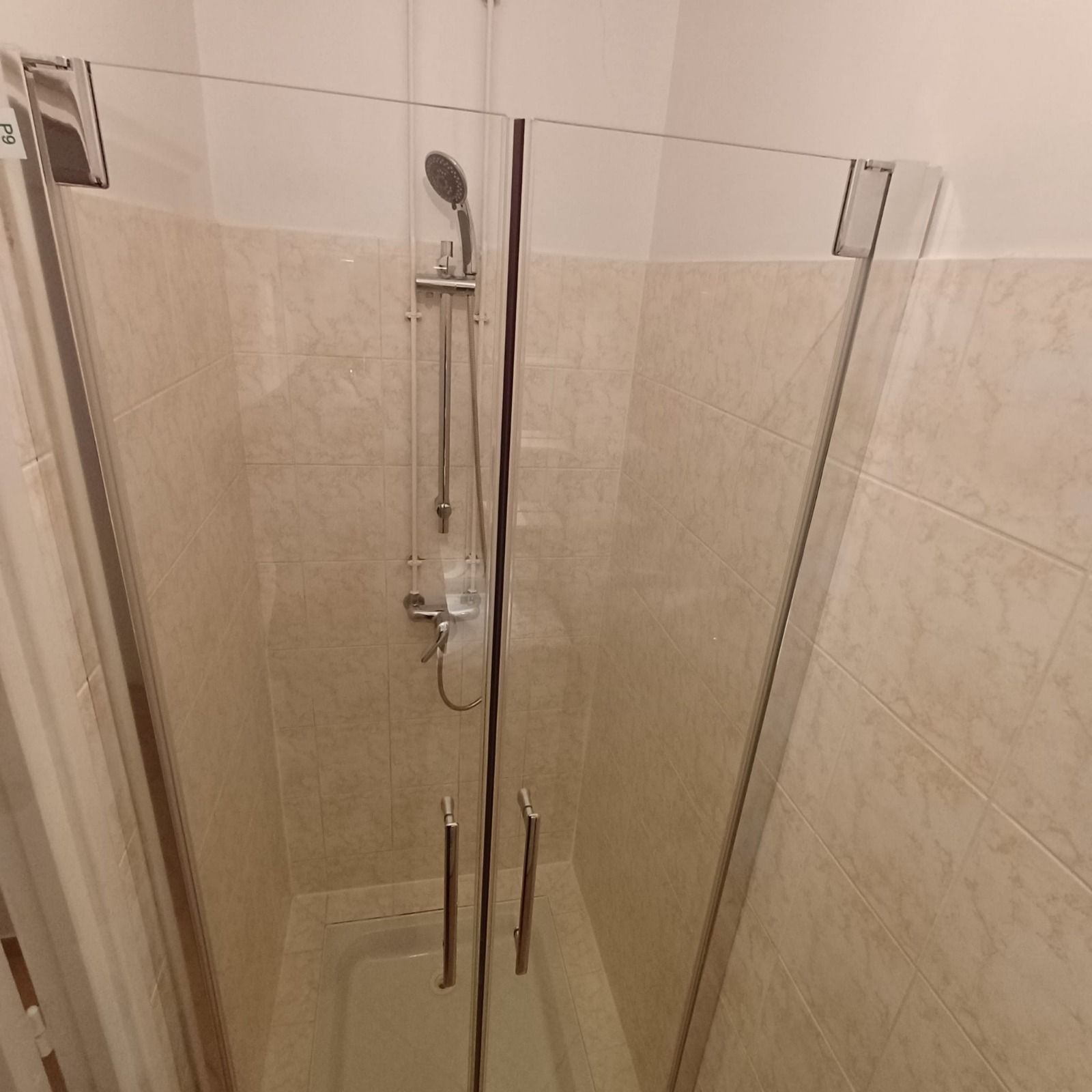 Une cabine de douche vitrée avec des murs carrelés beiges et une douchette à main sur une barre verticale.