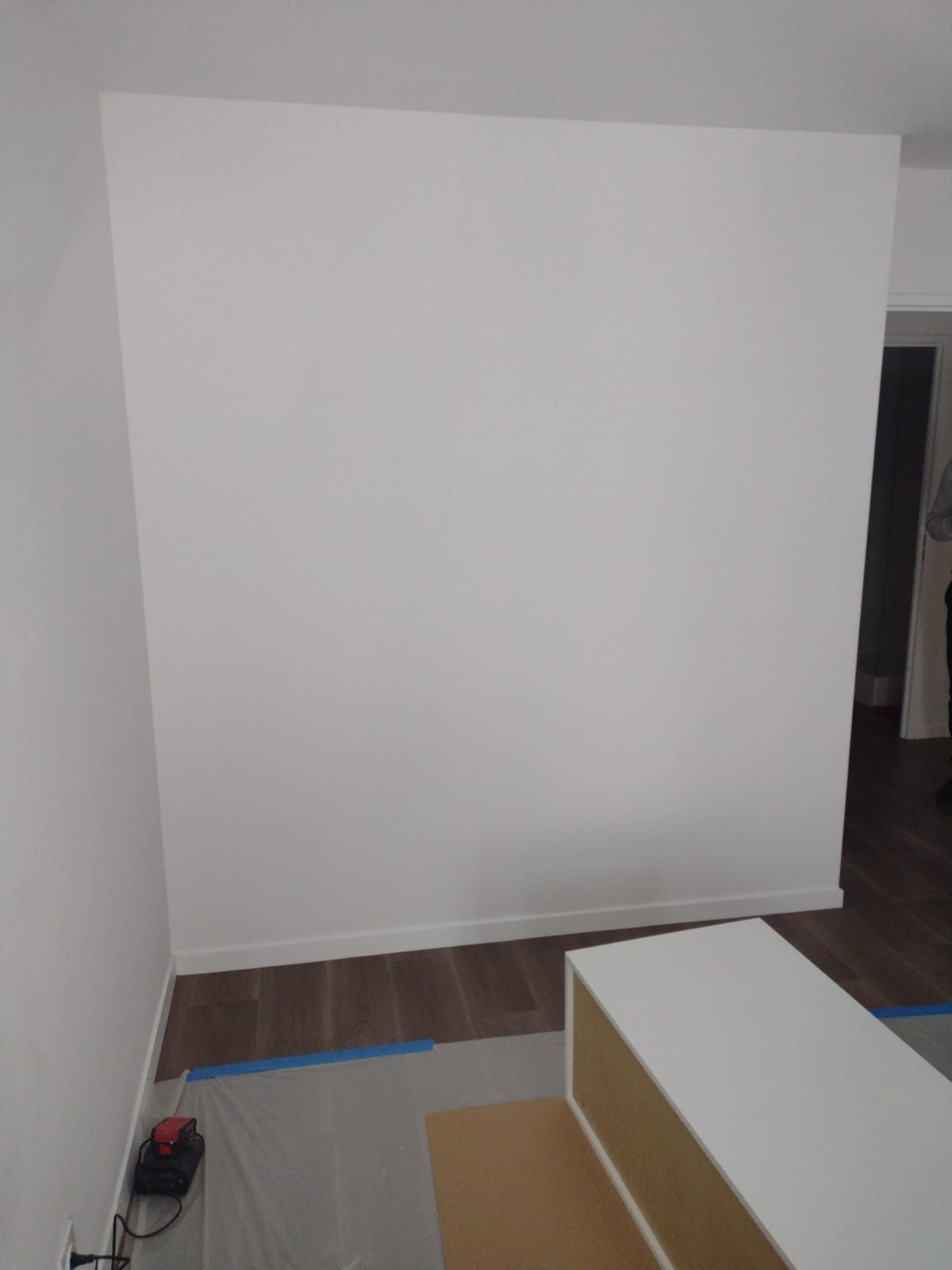 Un large mur blanc immaculé dans une pièce au parquet foncé, avec un coin de meubles blancs au premier plan.