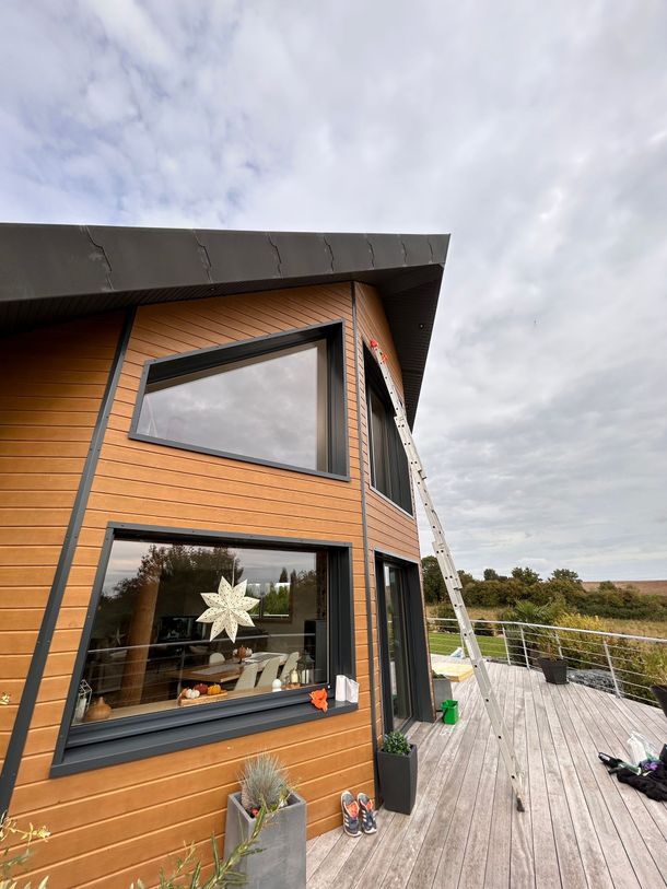 Maison en bois à ossature en A avec de grandes fenêtres, un toit noir et une cour enneigée sous un ciel nuageux.