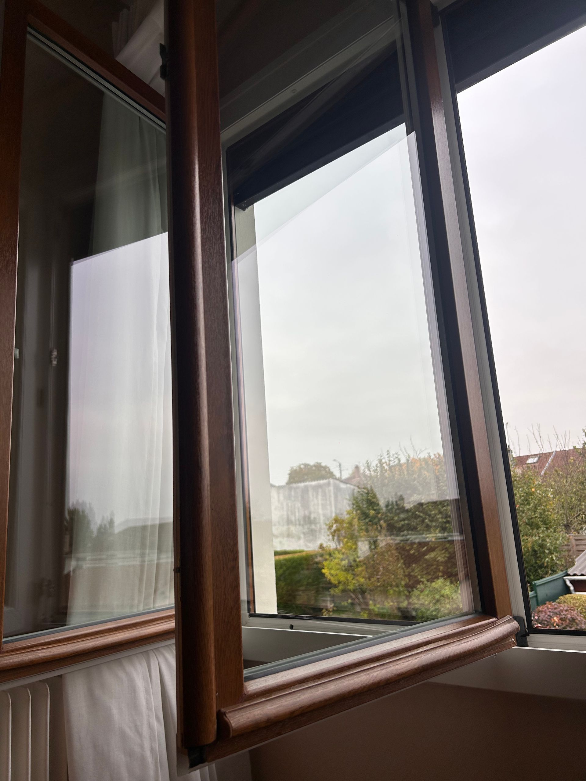 Fenêtre à cadre en bois ouverte sur une vue nuageuse d'arbres et de toits à l'extérieur.