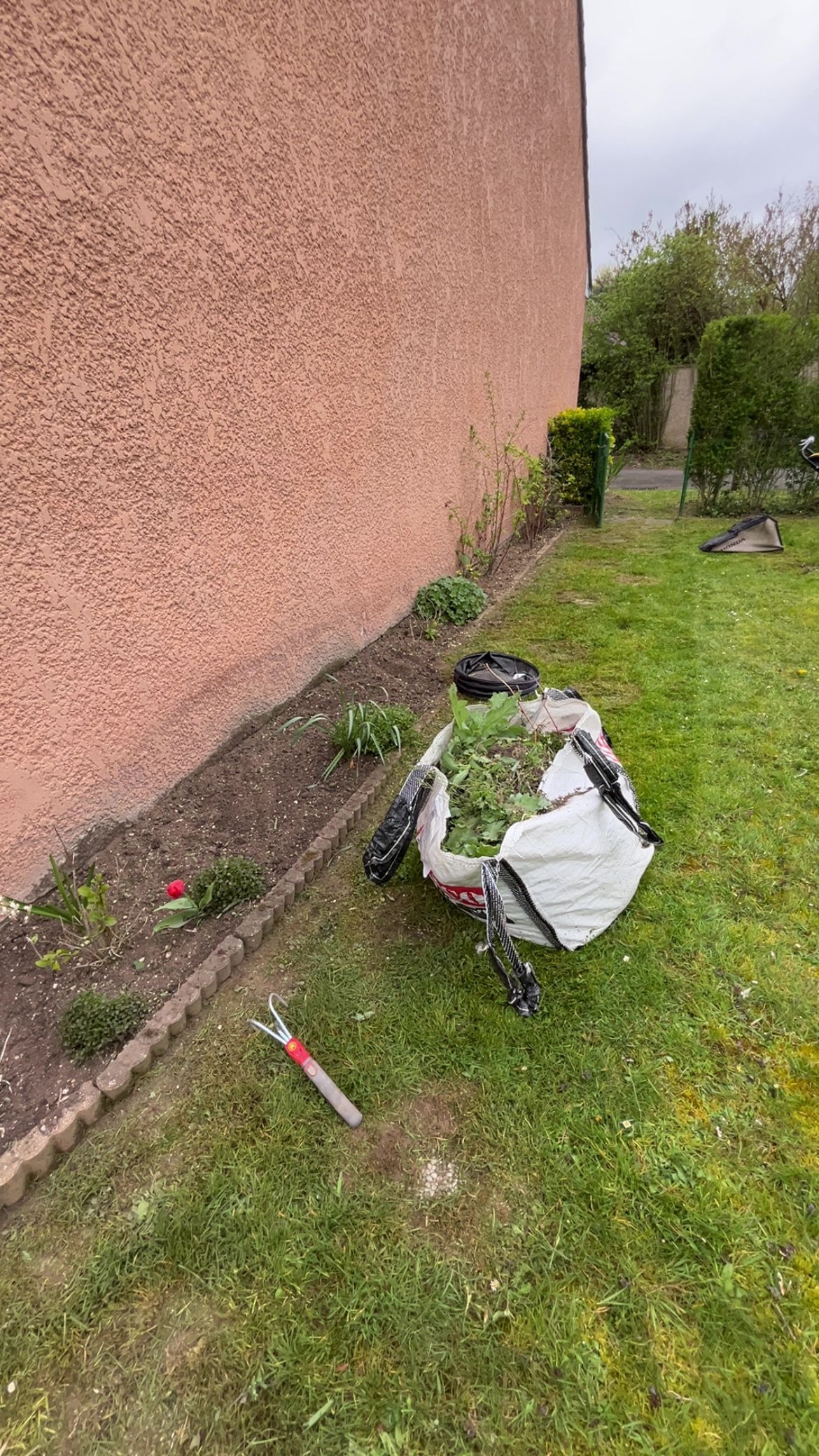 Des outils de jardin et un sac blanc gisent sur l'herbe à côté d'un mur en stuc rose.