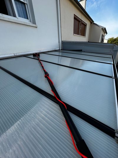 Des panneaux de toiture ondulés endommagés, traversés par une sangle rouge, jouxtent un bâtiment blanc.
