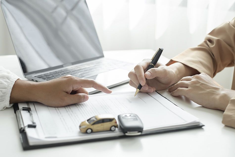Persona firmando un documento, llaves del coche y coche de juguete sobre el escritorio con una computadora portátil.