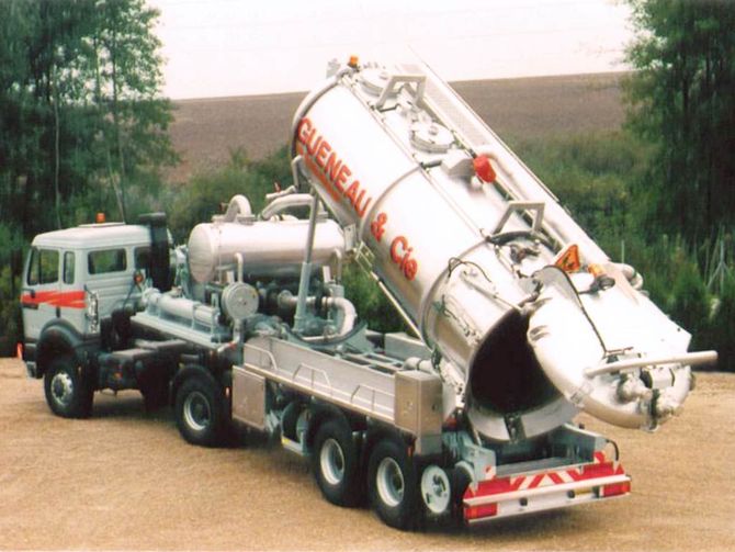 Gros camion aspirateur argenté, cuve levée, stationné à l'extérieur.