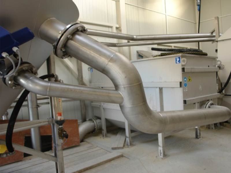 Des tuyaux en acier inoxydable sont raccordés à des machines industrielles dans une pièce aux murs blancs.