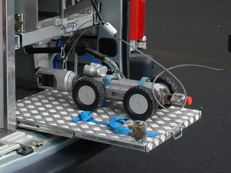 Un petit robot à roues, équipé de caméras et de câbles, est posé sur une plateforme métallique fixée à un véhicule.