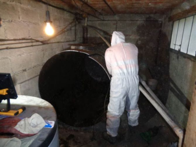 Une personne en combinaison de protection inspecte une grande cuve à eaux noires dans un sous-sol faiblement éclairé.