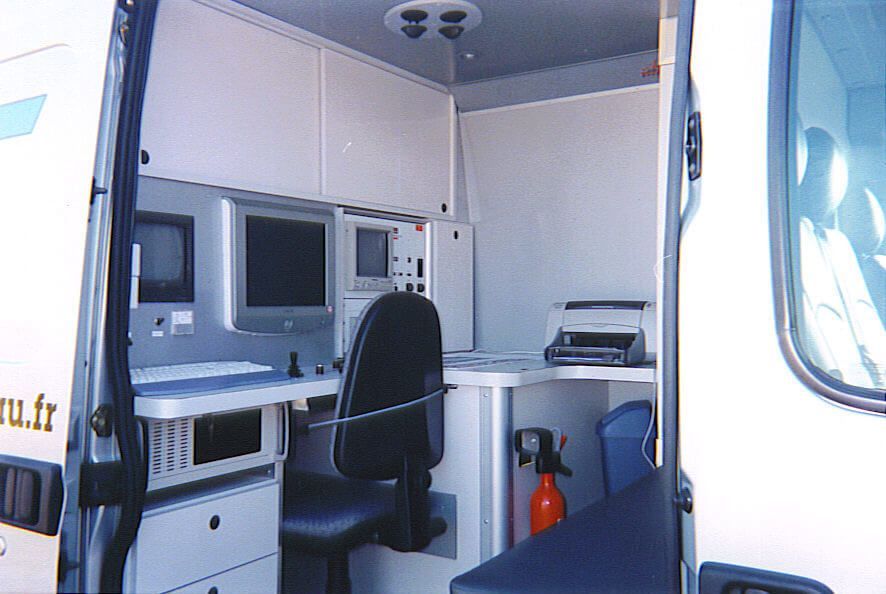 Intérieur d'un bureau mobile : murs blancs, écrans d'ordinateur, imprimante, chaise de bureau noire, extincteur, vu depuis la porte ouverte d'une camionnette.