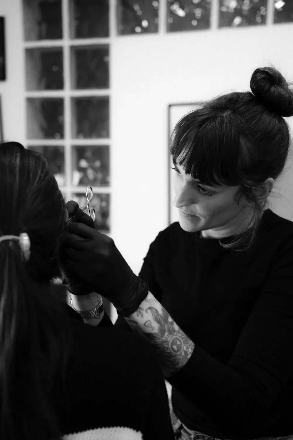 Persona con un tatuaje perforando la oreja de otra, ambas vestidas de negro. En un interior, cerca de una ventana.