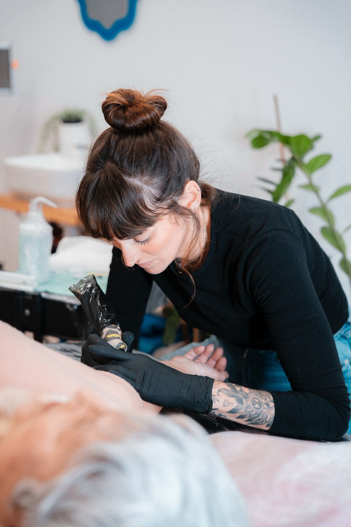 Mujer tatuando, inclinada sobre el brazo de un cliente. Ropa oscura y guantes. Máquina de tatuar visible. Estudio.